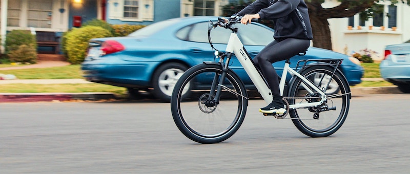 A woman wearing a helmet rides an electric bicycle along a street past a parked car