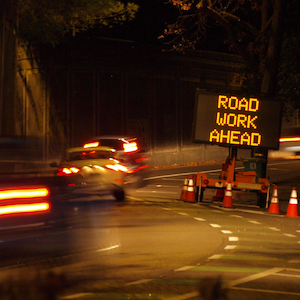 Cars driving past an illuminated sign saying 'Road Work Ahead' at night