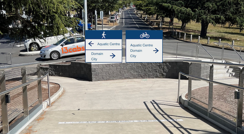 A photo taken coming off the western end of the Bridge of Remembrance. A pedestrian wayfinding sign has been added which shows Aquatic Centre to the left and Domain and City to the right. A cyclist wayfinding sign has been added which shows Aquatic Centre, Domain and City to the right.