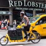A man rides a loaded yellow cargo bike down a New York street past a yellow taxi