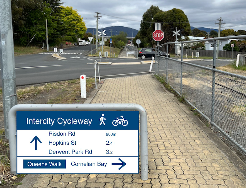 A photo taken on the Intercity Cycleway approaching the Queens Walk road crossing from the south. A wayfinding sign for pedestrians and cyclists has been added which shows Risdon Road, Hopkins Street and Derwent Park Road straight ahead via the Intercity Cycleway and Cornelian Bay via Queens Walk to the right.