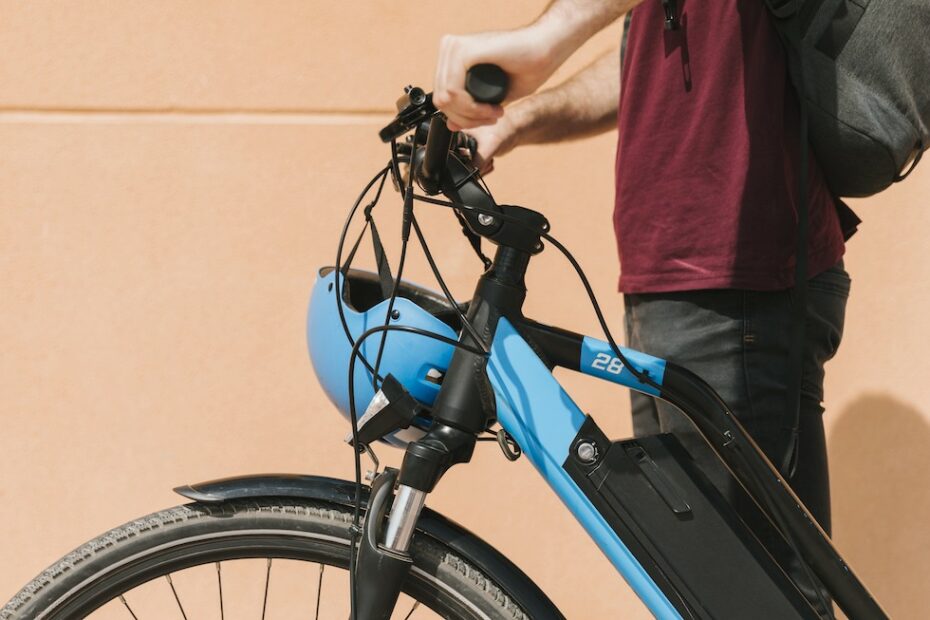 A photo showing the torso of a person wearing a backpack who is standing holding a blue e-bike.