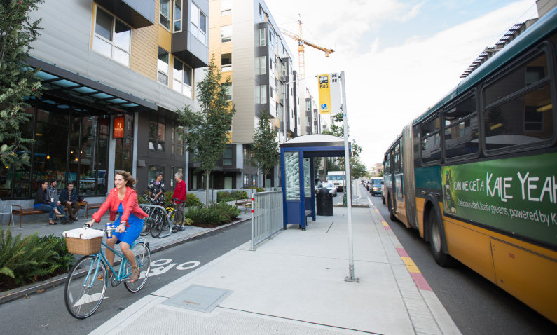 A bus stopped at a floating bus stop while a woman rides a bike along a bike lane between the bus stop and the footpath