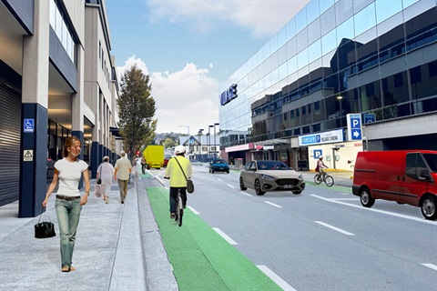 A person rides a bike along a green painted bike lane next to the kerb opposite the Village cinema complex. A second person rides in the opposite direction in a bike lane along the far side of the street. There's people walking on the footpath. Cars are being driven along the road. There is a red van parked on the far side of the road.