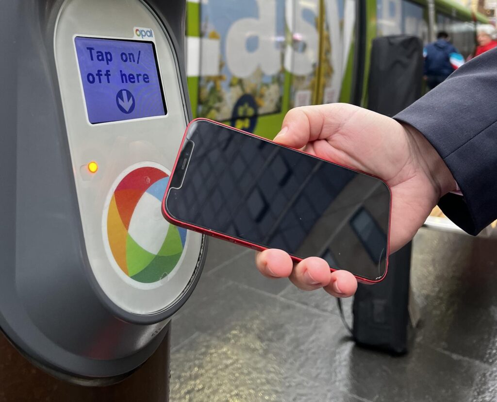 A hand holds a mobile phone towards a contactless payment reader which displays "Tap on/off here".