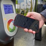 A hand holds a mobile phone towards a contactless payment reader which displays "Tap on/off here".