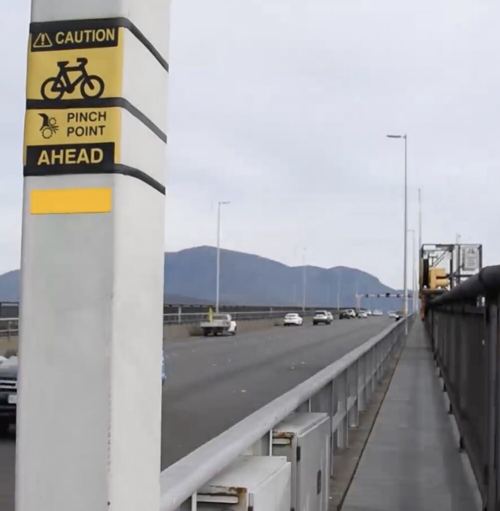 A photo of a very narrow path next to a roadway on a bridge. There's a gantry on the fence at one point making it even narrow. A sign reads "Caution, pinch point ahead". There's several cars. There's no people walking or riding.