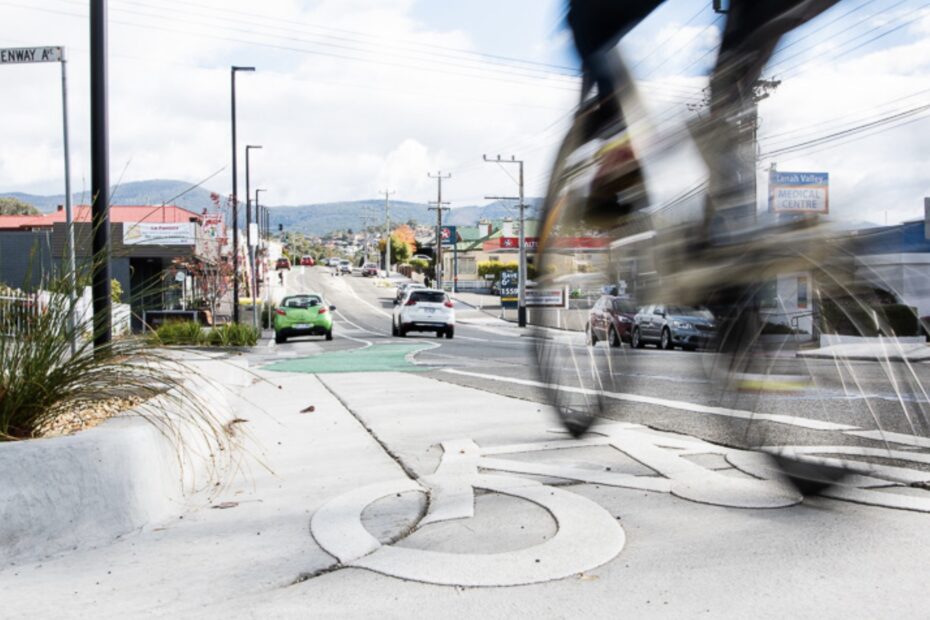 A photo showing a bike lane on Augusta Road Lenah Valley with a motion blurred person riding a bike passing in the foreground. There's moving and parked cars in the background.