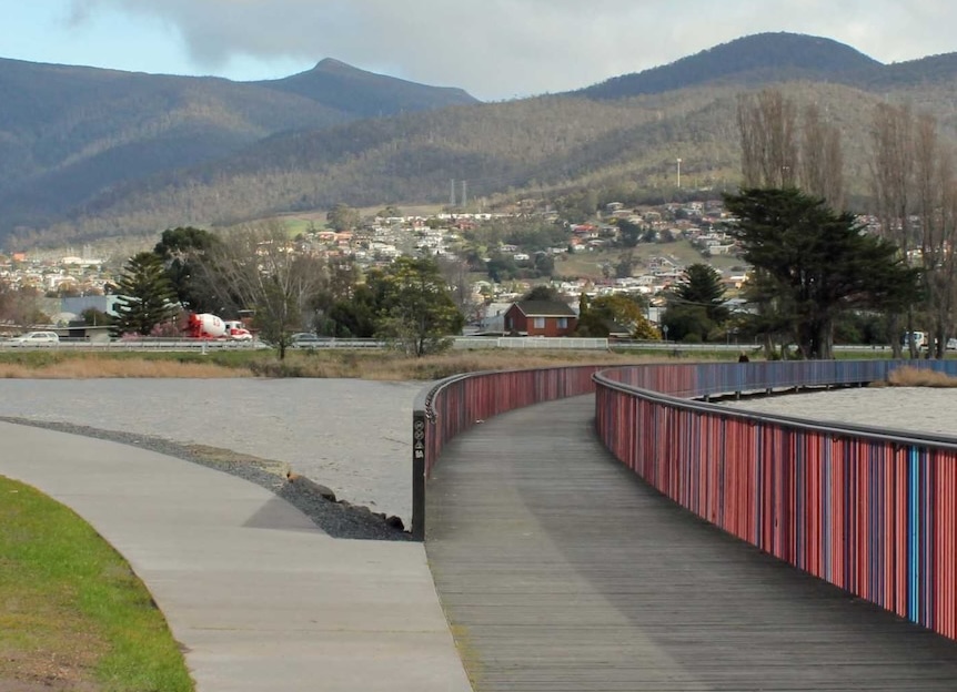 A photo of a walking and riding path going over a colourful bridge over a body of water. There's forested hills in the background and a cloudy sky.