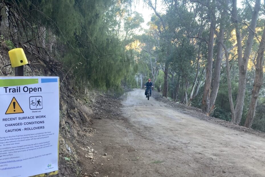 A person riding a bike up the Bumpy Track fire trail in South Hobart. It is now a relatively smooth gravel fire trail through the bush. A temporary City of Hobart sign reads: "Trail Open. Recent surface work. Changed conditions. Caution - rollovers.